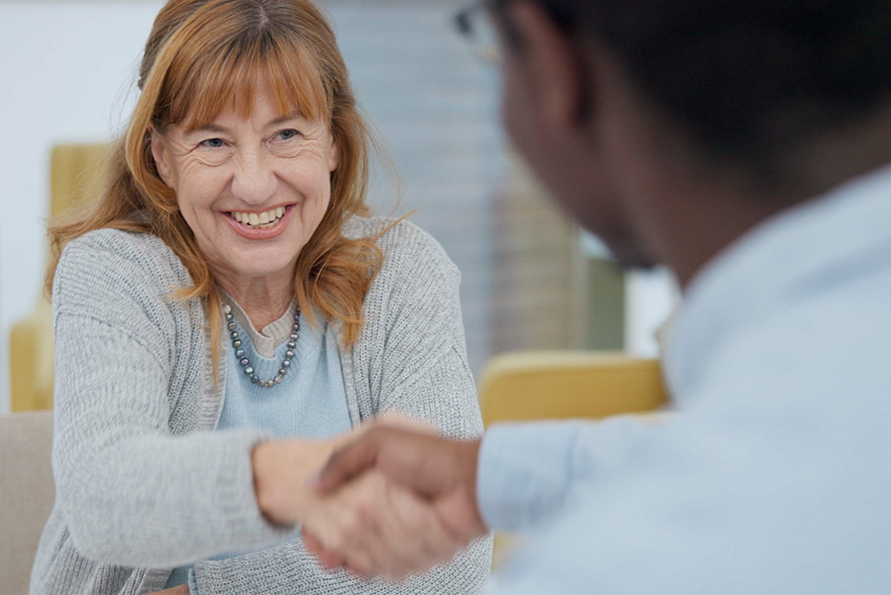 image_2 A smiling woman with light brown hair shakes hands with a man across a table in an office.